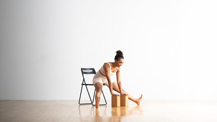 Woman practices Tittibhasana, Firefly Pose sitting in a black folding chair. She leans forward and places her hands on two cork yoga blocks. She is sitting against a white wall.