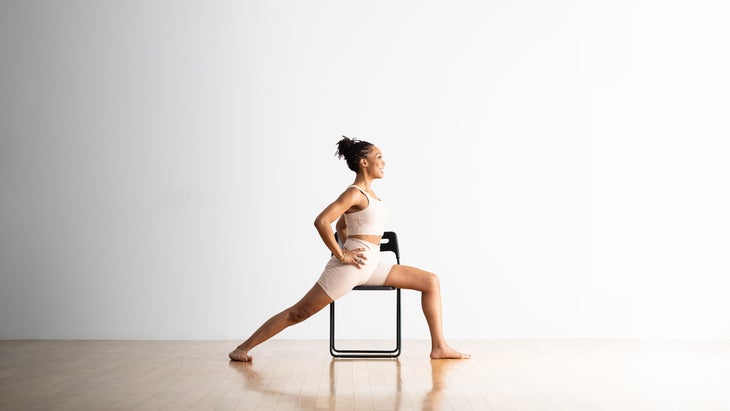 Woman practices Warrior I pose in a chair. She sits astride the chair facing right, with one leg behind the others. She is wearing a light yoga plans and top, against a white background
