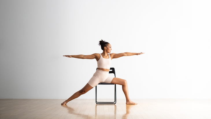 Woman practices Warrior II pose in a chair. She sits astride the black chair facing right with her arms extended at shoulder height. She is wearing a light yoga plans and top, against a white background