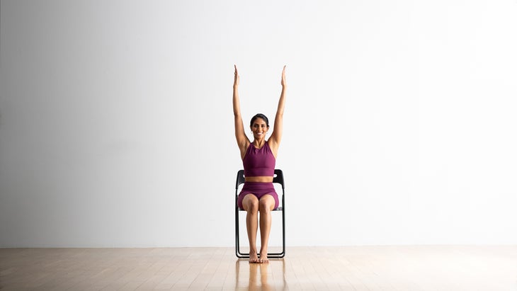 Person extends her arms up to practice Urdhva Hastsana in a black folding chair against a white background. She is wering deep pink tank and shorts.