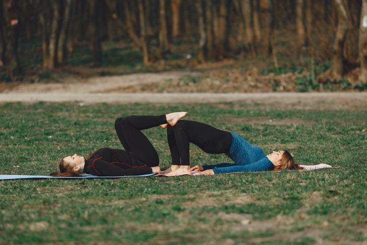 Two women in dark yoga clothes practice a couples variation of bridge pose. They are practicing outside on grass.