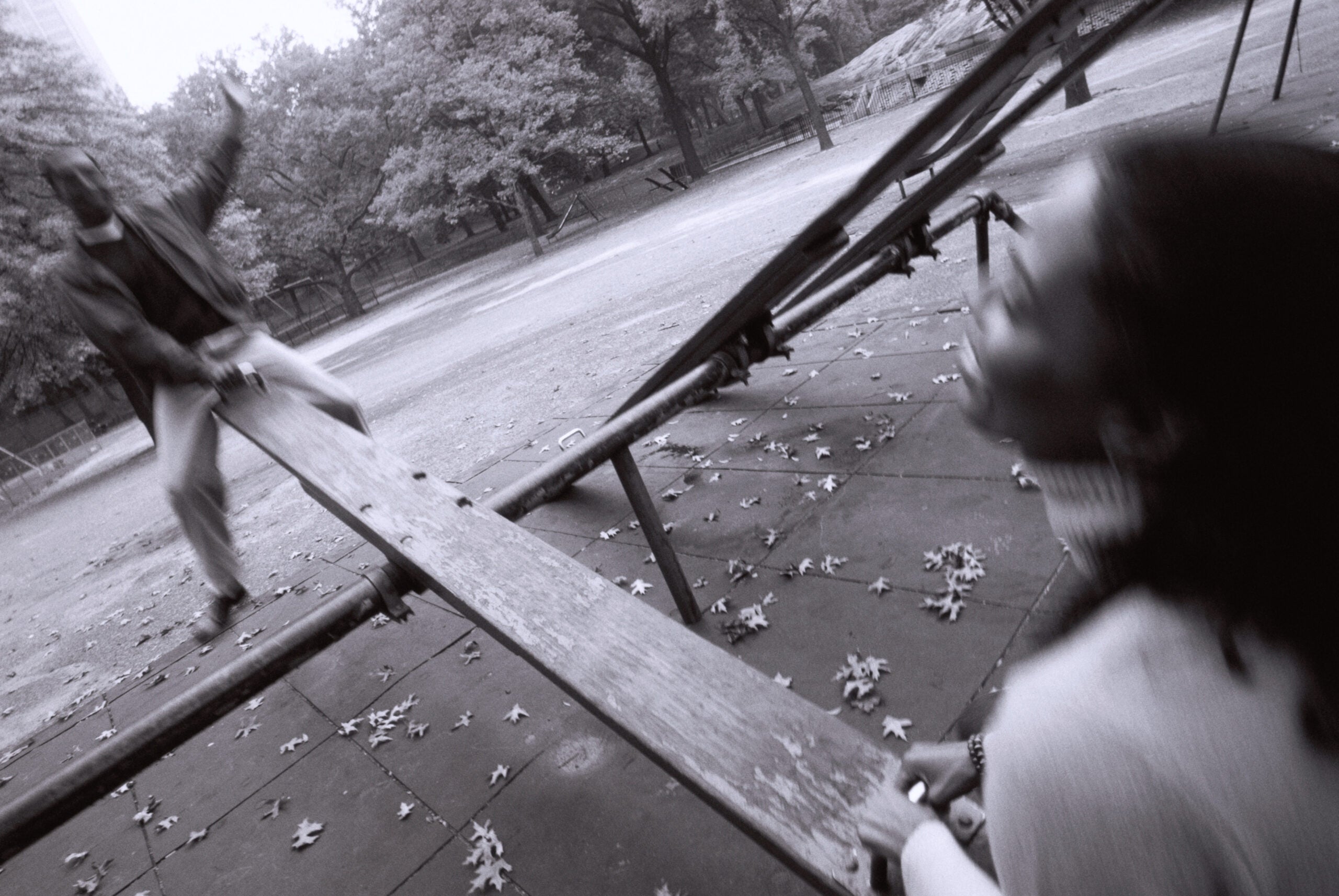 A blurred black and white image of a woman and a man on a see saw. There are trees in the background.
