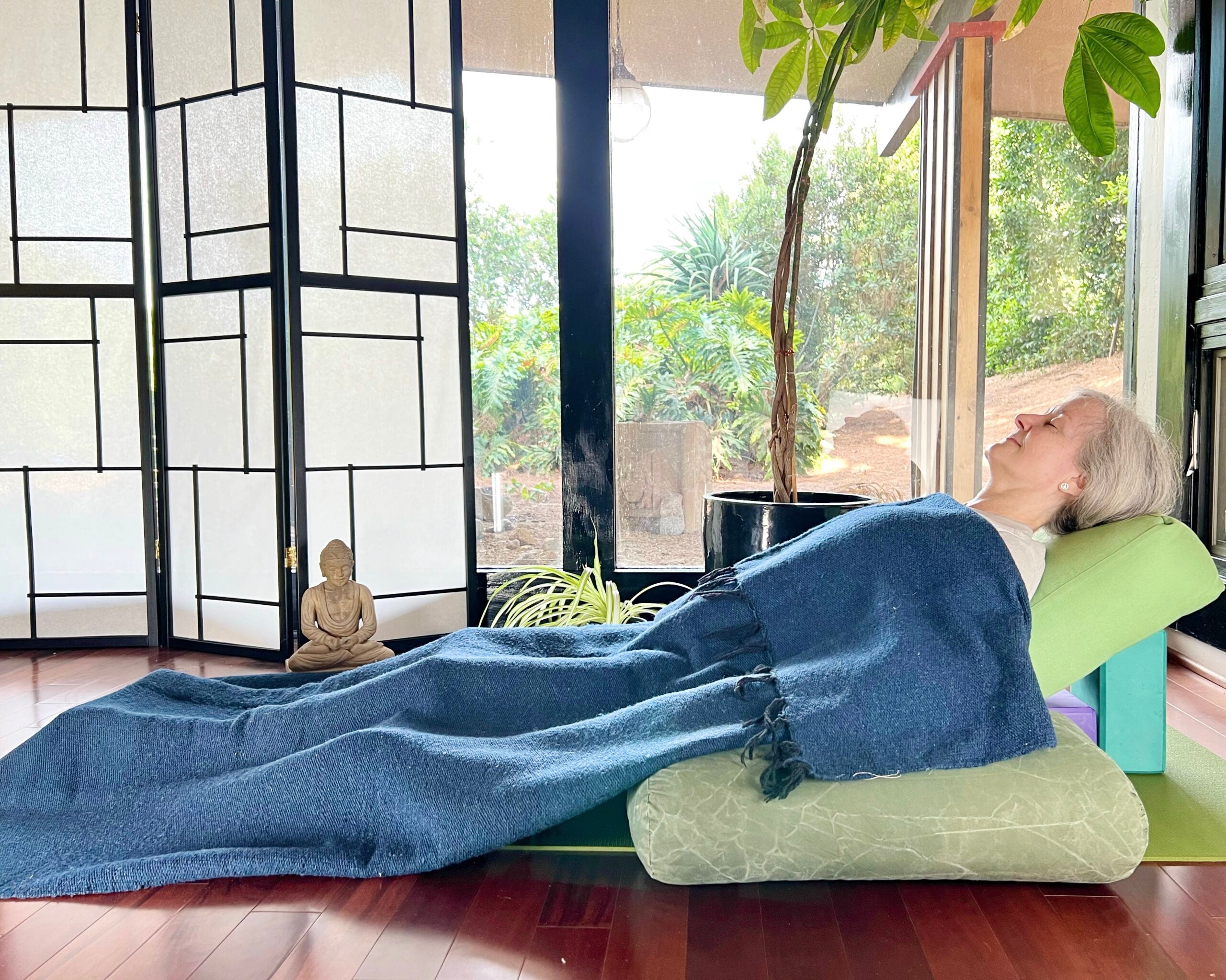 A woman with white hair practices Savasana, Corpse Pose. She is lying on a green mat on a brown wood floor, wearing brown pants and a light brown shirt. In the background are floor-to-ceiling windows with a view of trees. A shoji screen and a small Buddha statue are on the left and a tall plant is behind her. She is lying on a bolster that is propped up by blocks. A blue blanket is draped over her body.
