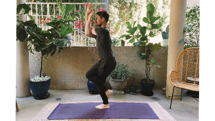 Man standing on a yoga mat with his right leg crossed over his left leg and his arms intertwined in Eagle Pose