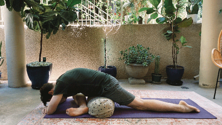 Man stretching his hips by draping his body over a bolster on his yoga mat in Pigeon Pose