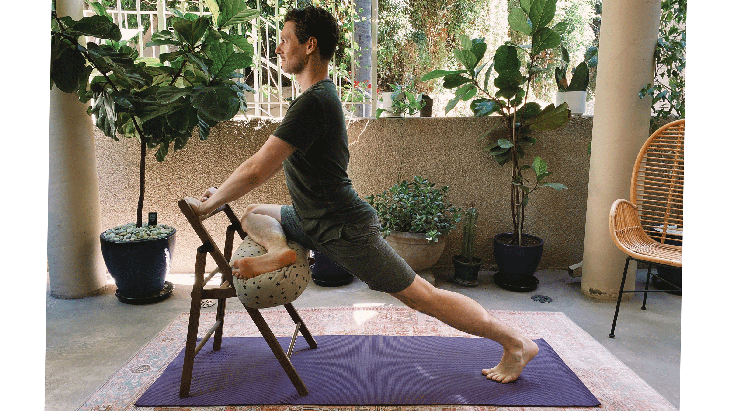 Man practicing PIgeon Pose in yoga with a bolster on a chair and his front knee bent and on the bolster