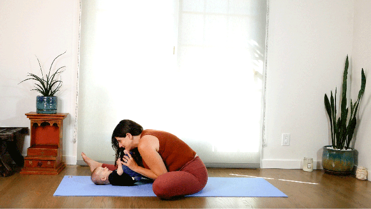 Woman sitting on a yoga mat and leaning forward toward her baby while one knee is bent and the other is straight in a yoga pose