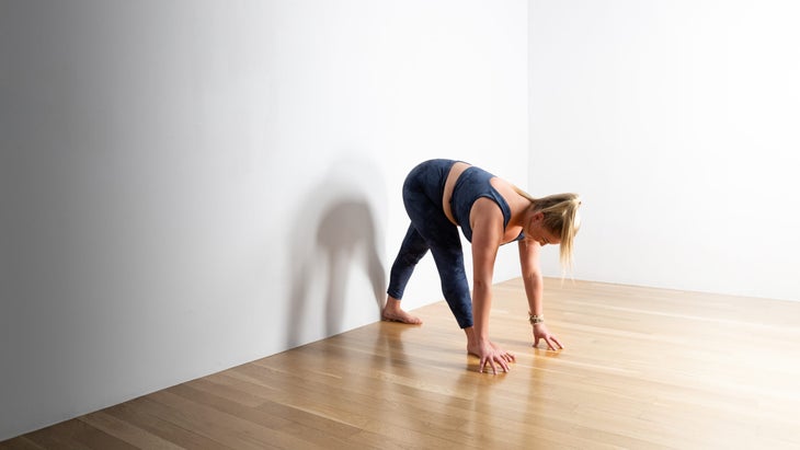 A woman in blue tights practices Pyramid pose with her left foot against a white wall