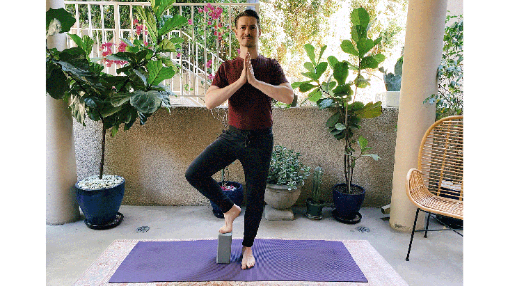 Man standing on a yoga mat balancing on one leg in Tree Pose with his right foot on a block for balance
