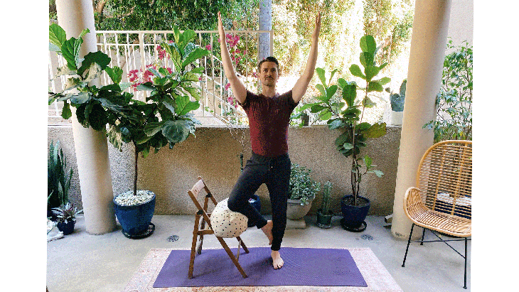 Man standing on a yoga mat balancing on one leg in Tree Pose with his right knee bent and resting against a chair for steadiness
