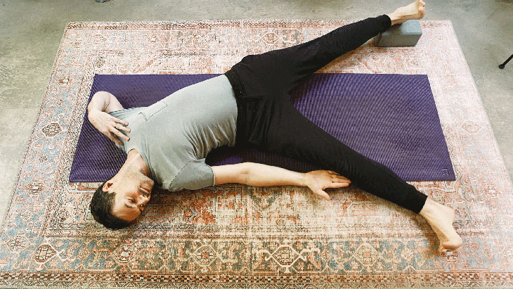 Man lying on his back on a yoga mat with his right hand reaching for his right shin in Triangle Pose