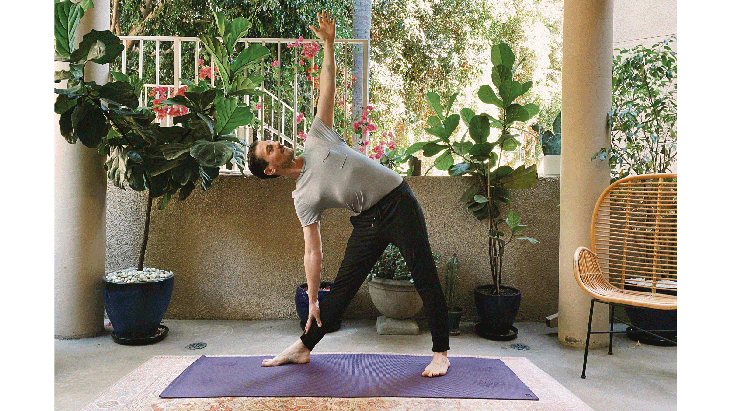 Man standing on a yoga mat with his legs in Triangle Pose and his right arm reaching to his shin and his left arm reaching to the ceiling