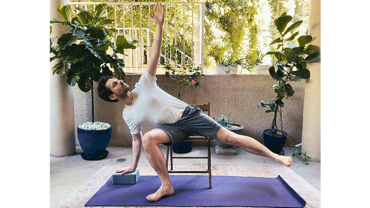 Man seated on a chair with his right knee bent and his right hand on the yoga mat with his left leg lifted in Half Moon Pose, a yoga posture