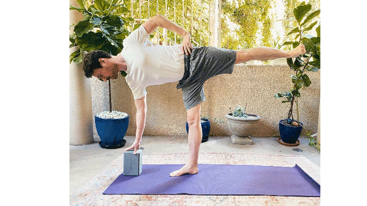 Man standing on a yoga mat balancing on one leg with his right hand on a block and his left hand on his hip. He's on a patio with plants in the background.