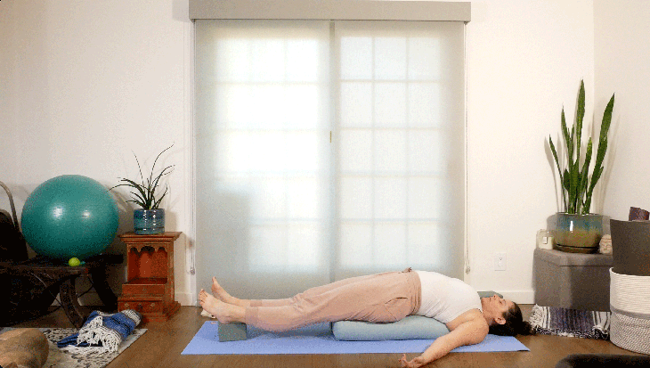 Woman lying on her yoga mat with a bolster beneath her back and a block beneath her feet resting in Bridge Pose