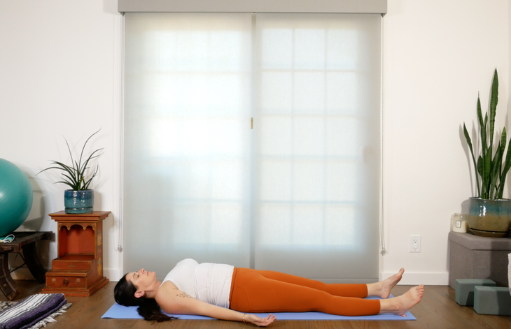 Woman lying on her back in Savasana with her legs, body, and arms relaxing on the mat and her eyes closed