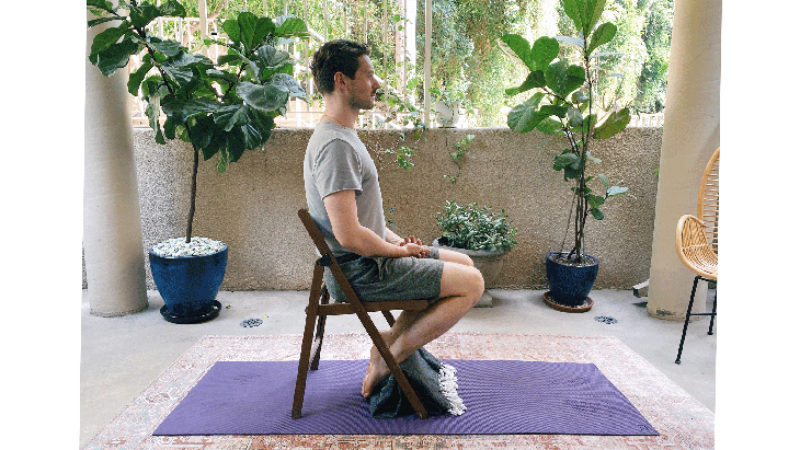 Man sitting on a chair with his feet tucked behind the front run and his hands in his lap. He's practicing a variation of the yoga posture Virasana (Hero Pose).