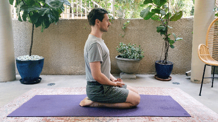 Man kneeling on a yoga mat with his heels beneath him. His hands are resting in his lap and his shoulders are over his hips.