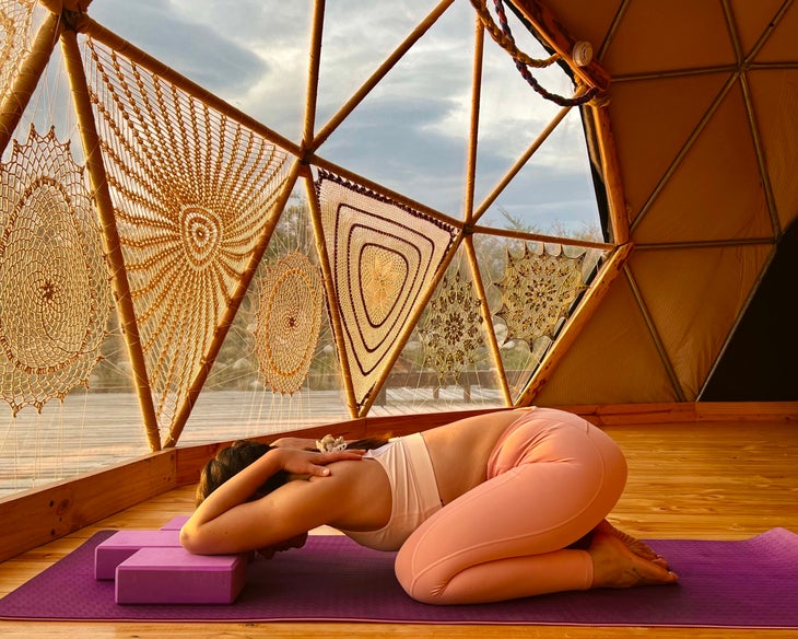 Woman with dark braid practices yoga--Balasana (Child’s Pose with bent forearms)-- in a YogaDome, a geodesic dome in Patagonia. She is wearing peach colored tights and a light colored sports bra, and practices on a magenta colored mat. The room has wood floors and you can see crocheted covers on the triangular shaped windows. A view of the sky can be seen through the windows.