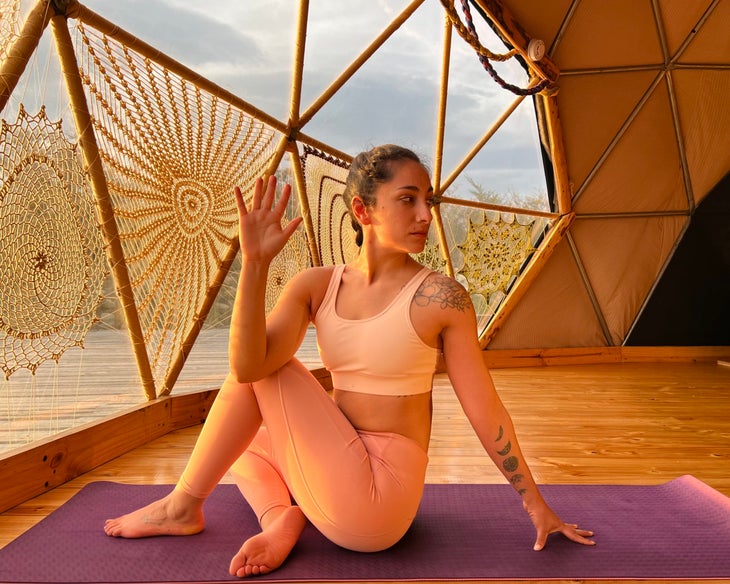 Woman with dark braid practices Ardha Matsyendrasana (Lord of the Fishes) in a YogaDome, a geodesic dome in Patagonia. She is wearing peach colored tights and a light colored sports bra, and practices on a magenta colored mat. The room has wood floors and you can see crocheted covers on the triangular shaped windows. A view of the sky can be seen through the windows.