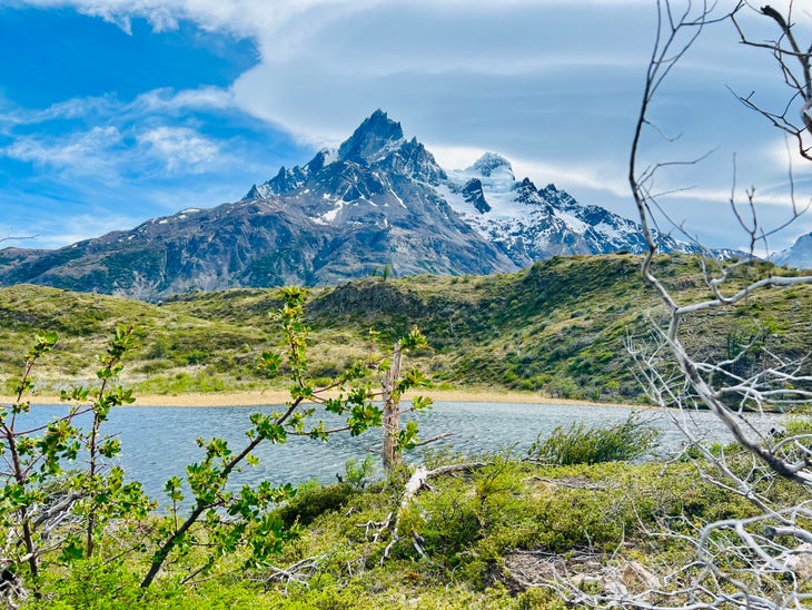 The snow-dusted mountains of Patagonia against the blue sky. In the foreground are brushy green hills with a blue river running through.