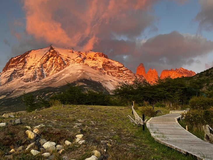 The sun shines on snowy mountains in Patagonia. In the foreground there is rocky green terrain and a wooden footpath.