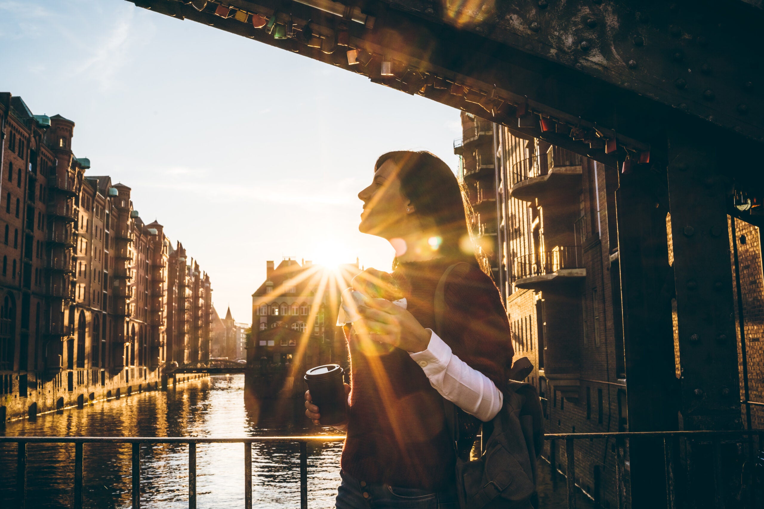 A young woman stands on a bridge in a historic part of Hamburg, Germany. The sun is shining behind her, placing her in silhouette. Behind her there are buildings along the river. She is holding a cup in her hand.