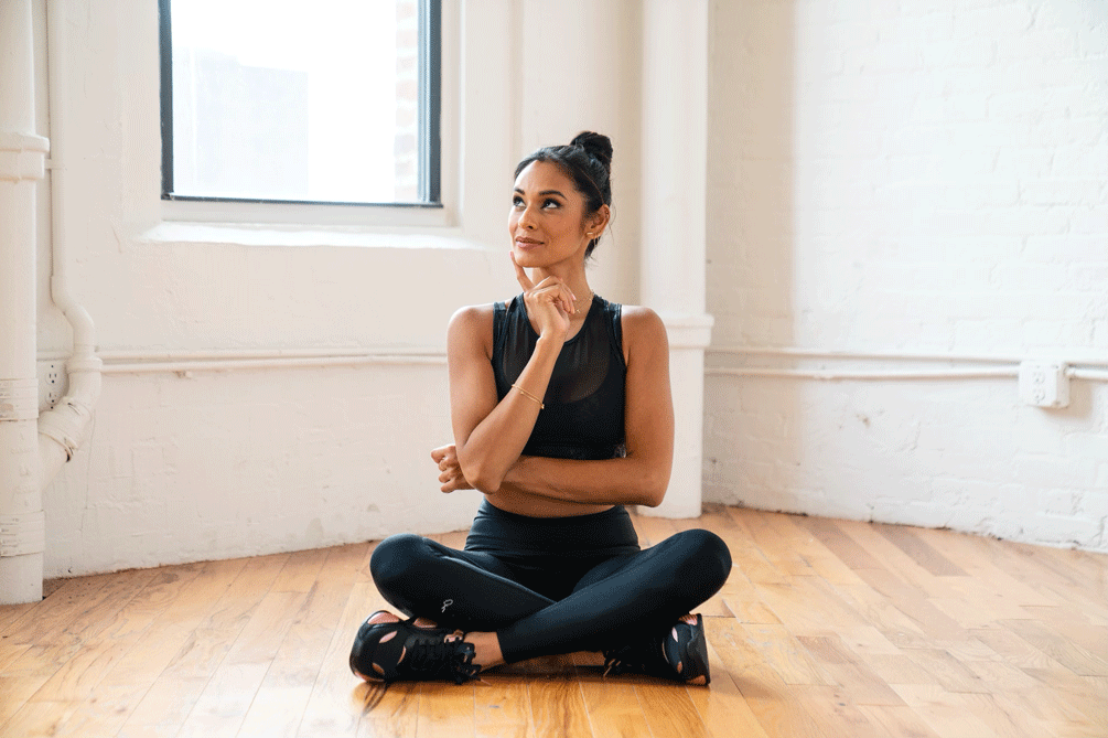 Yoga and meditation Aditi Shah of Peloton sits on a hardwood floor in a yoga studio with windows behind her as she contemplates meditation myths