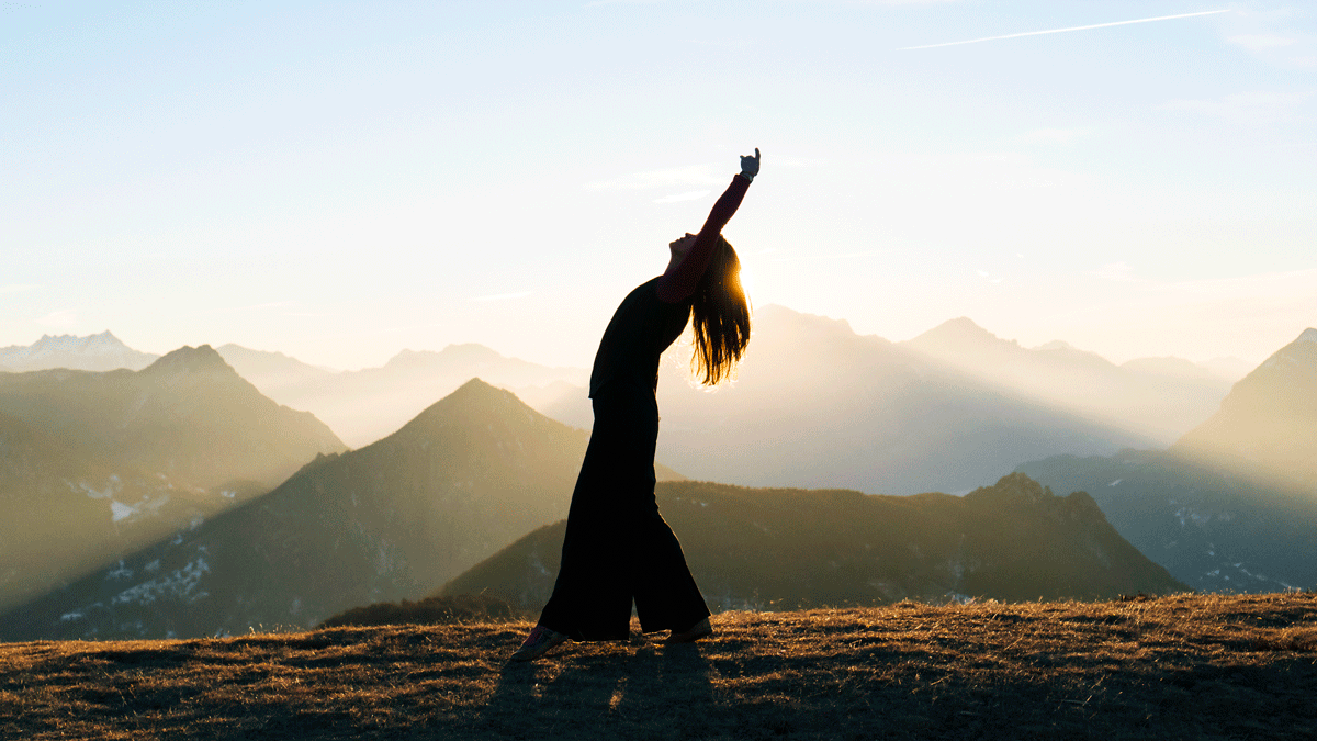 Woman standing outside at sunrise with mountains in the background and she is reaching her arms above her head and surrendering to her weekly astrology forecast