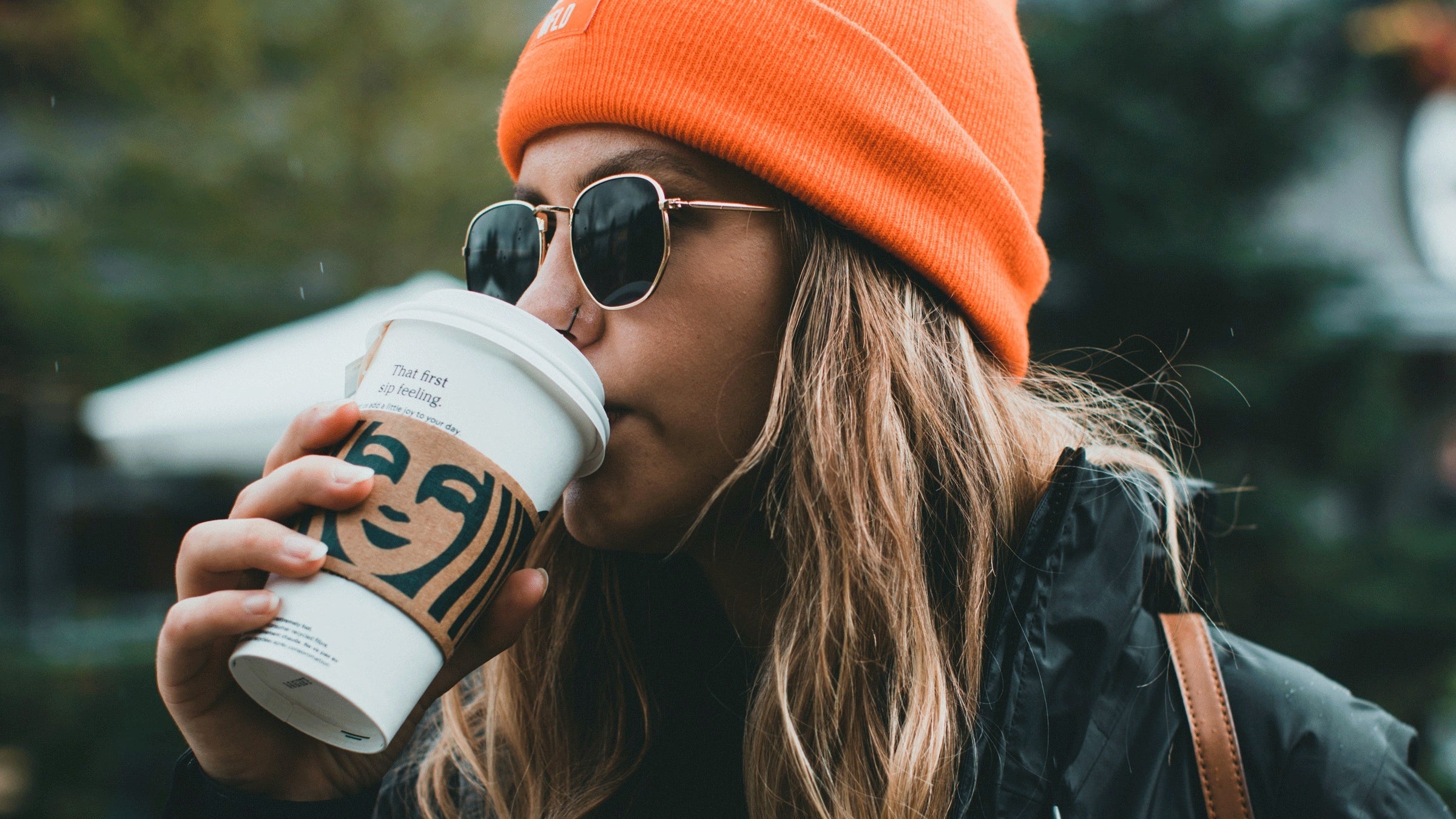 Woman outside with an orange stocking hat and sunglasses and puffer coat drinking coffee from Starbucks Star Signs