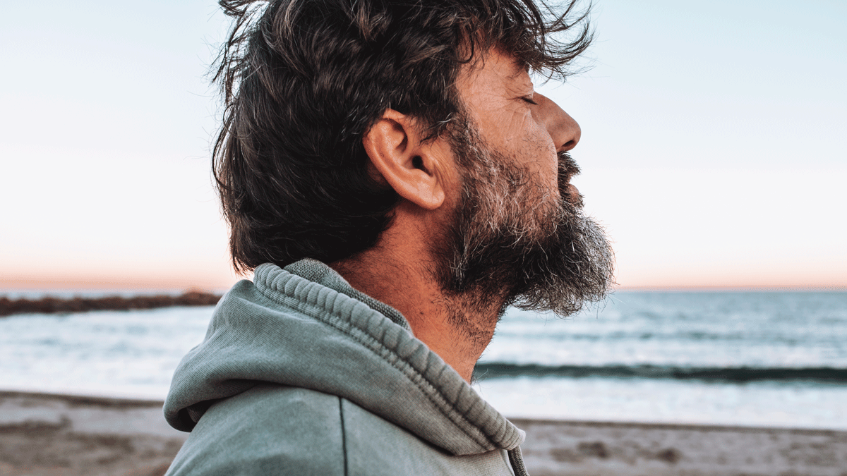Older man with salt and pepper hair and beard standing at the beach with eyes shut