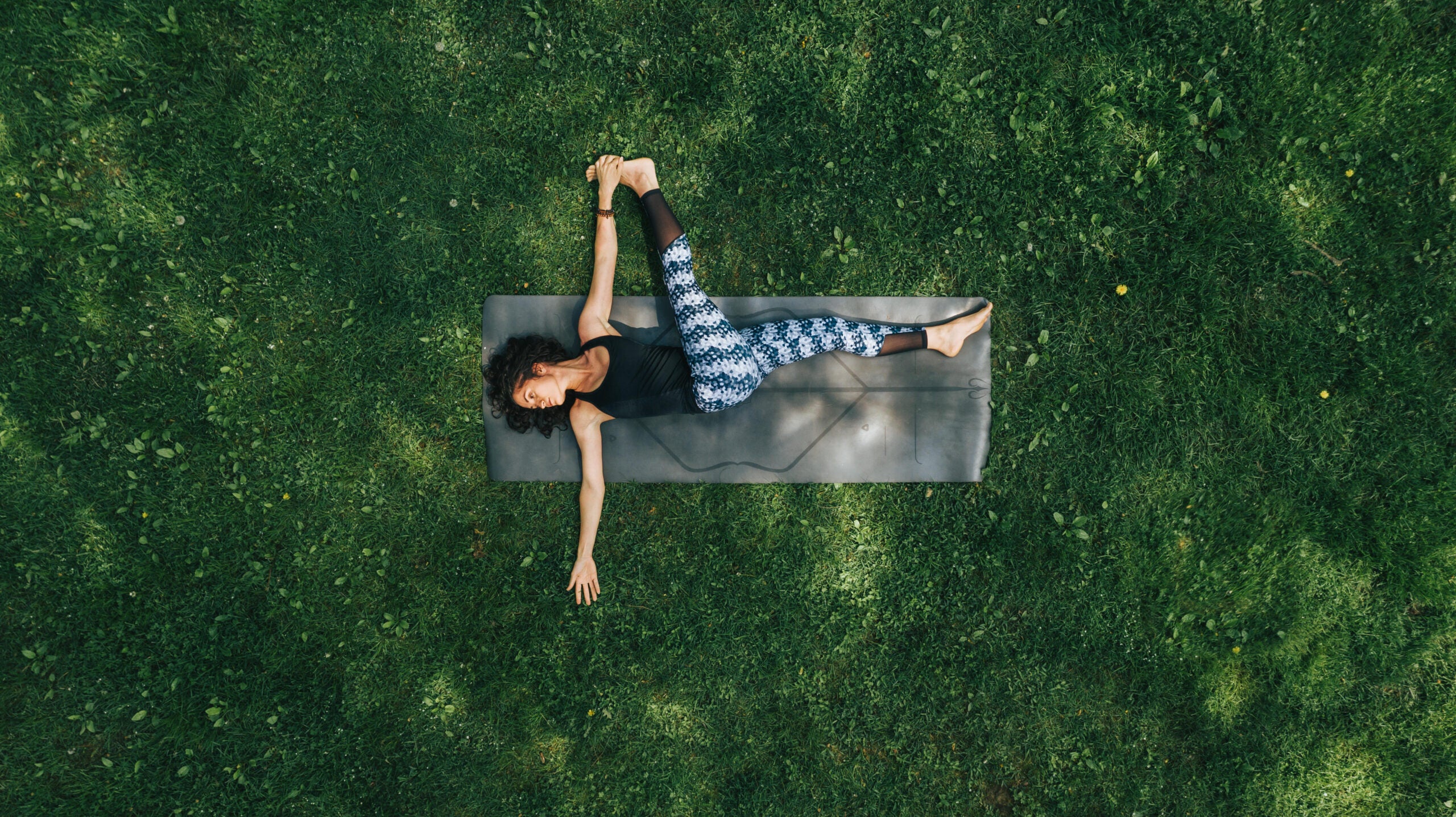 An aerial shot of a woman lying in an expanse of green grass. She is lying on a gray yoga mat doing a supine twist with her legs extended.