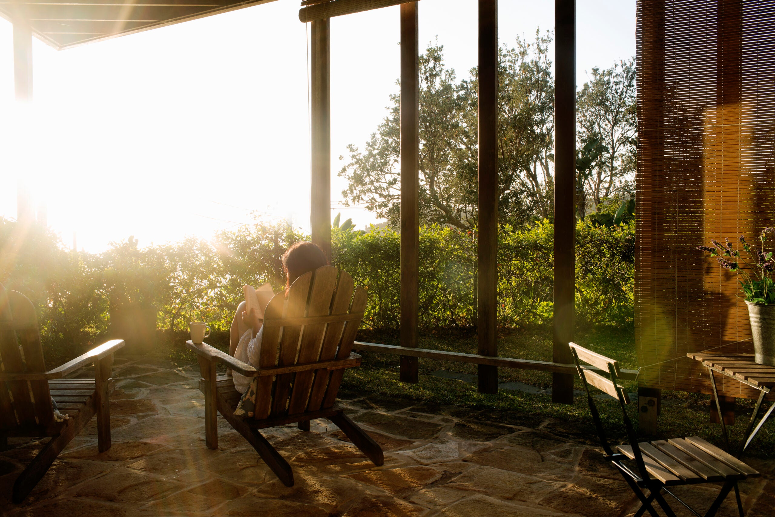 Person is sitting in an Adirondack chair on a porch with a stone floor. It is sunrise or sunset and light is streaming in from the left side of the frame. In the background there are trees and a table and chair behind the person.