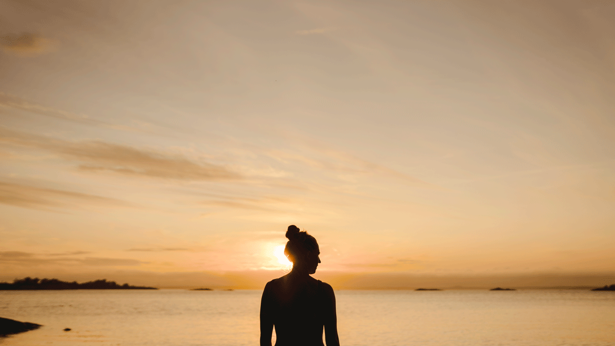 Woman standing on a beach at sunset pondering the weekly astrology forecast