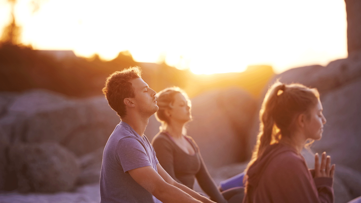 A man and two women sitting on the beach at sunrise meditating during a yoga retreat