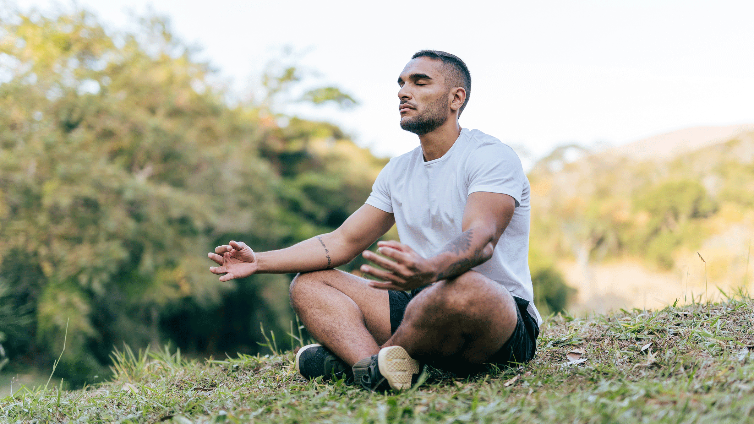 Young guy with tattoos sitting crosslegged outside in a park meditating while he recovers from an injury