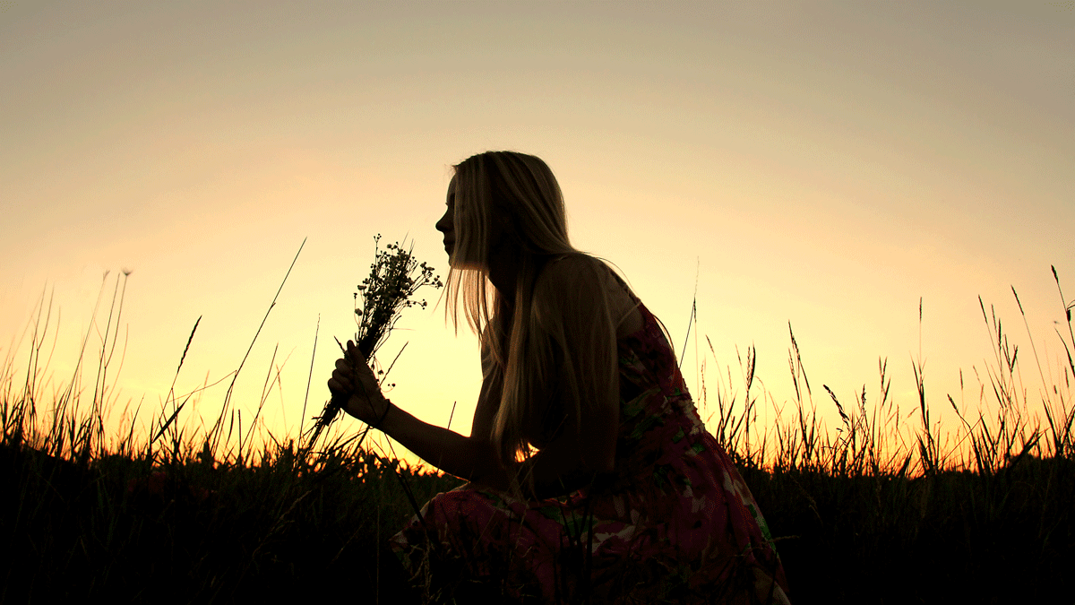 Woman sitting outside at dusk in a grassy field leaning forward and smelling a bunch of wildflowers under the new Moon September 2023, which is in Virgo