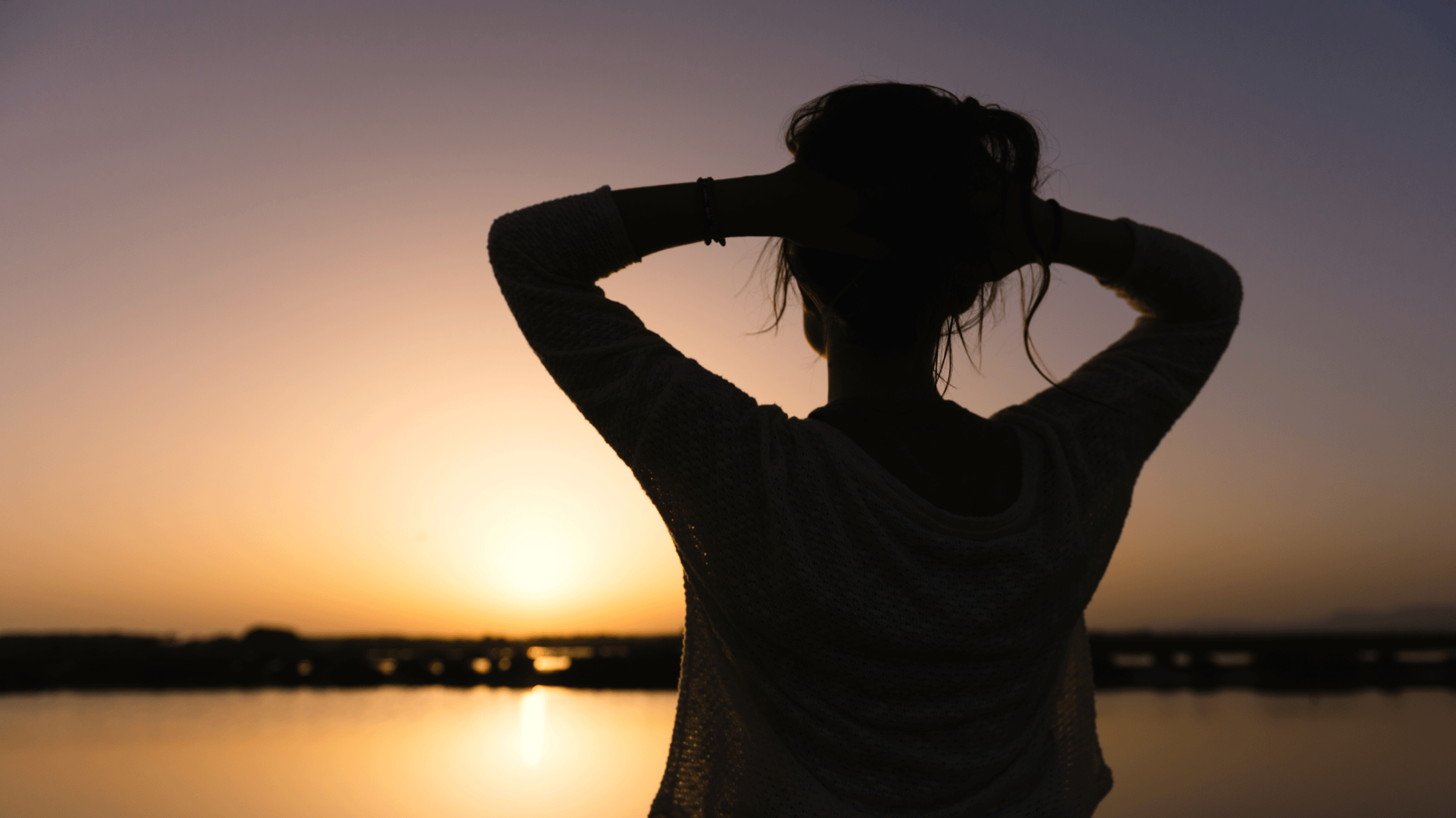Woman standing outside overlooking a bay at dusk while contemplating the weekly astrology forecast.