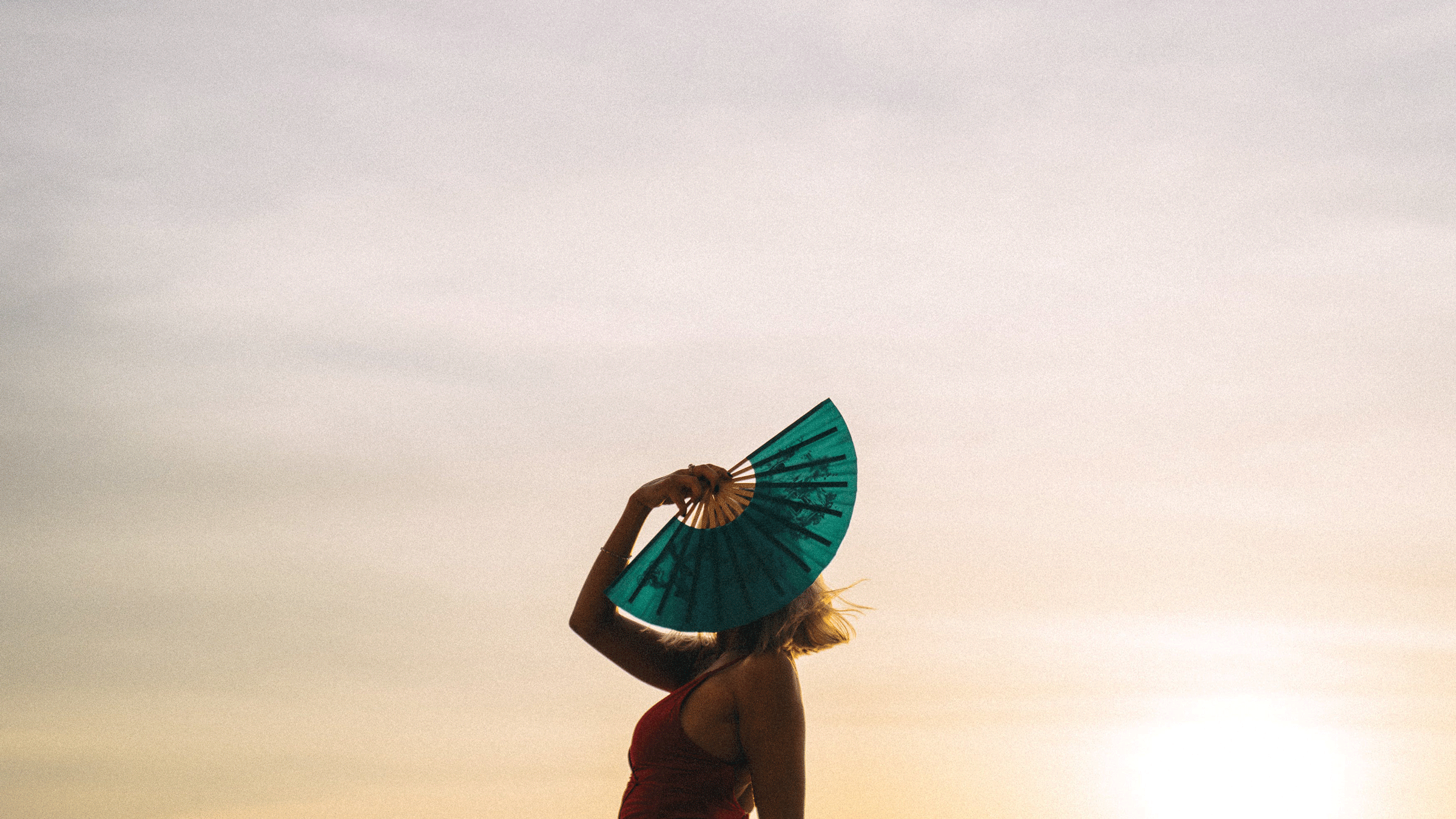 Woman standing outside just before dusk holding a paper fan in front of her face as she contemplates her future and the weekly astrology