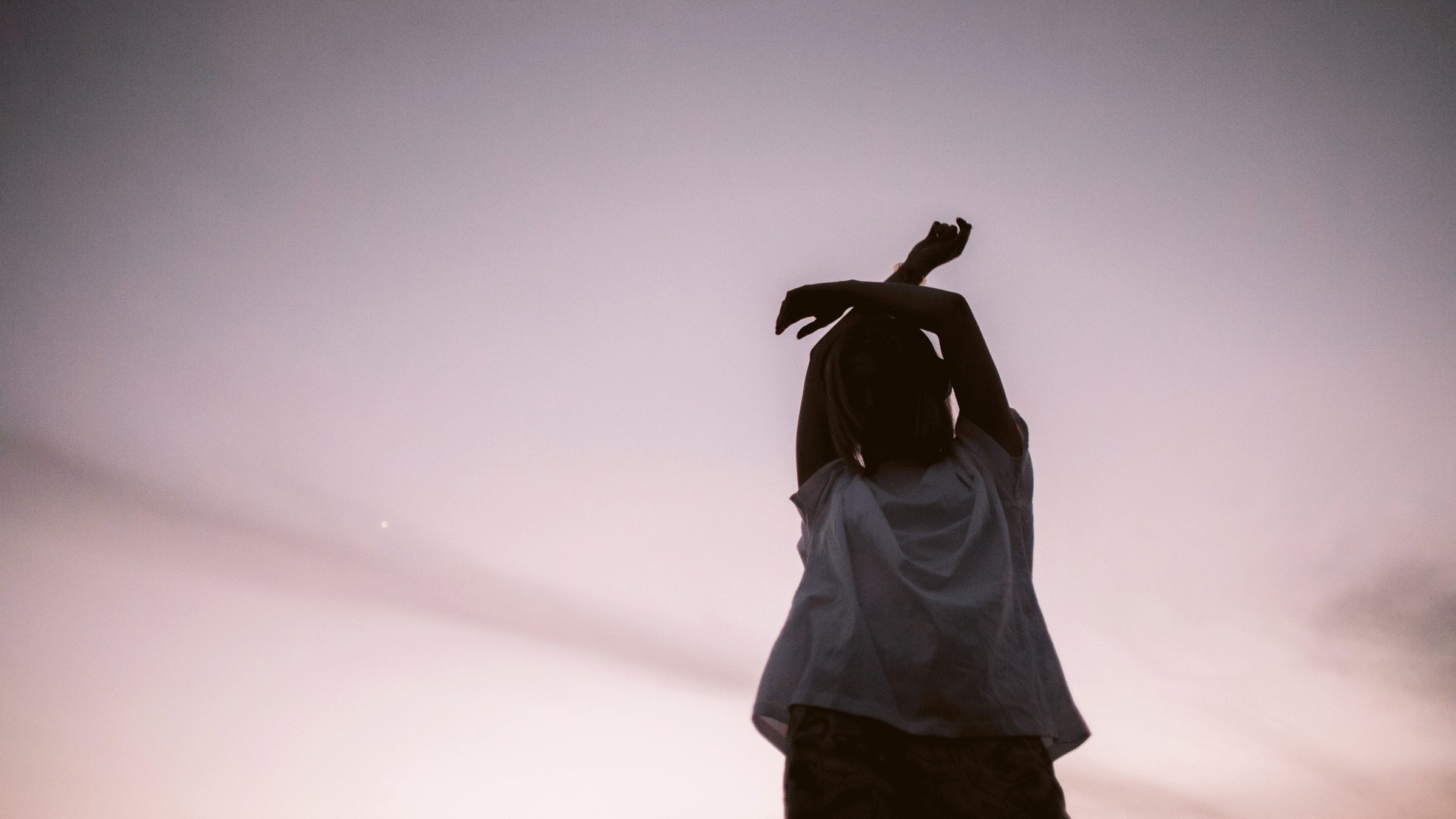 Woman standing outside at dusk with the mauve light behind her as she has her arms above her head. She's wearing a t-shirt and faded jeans.