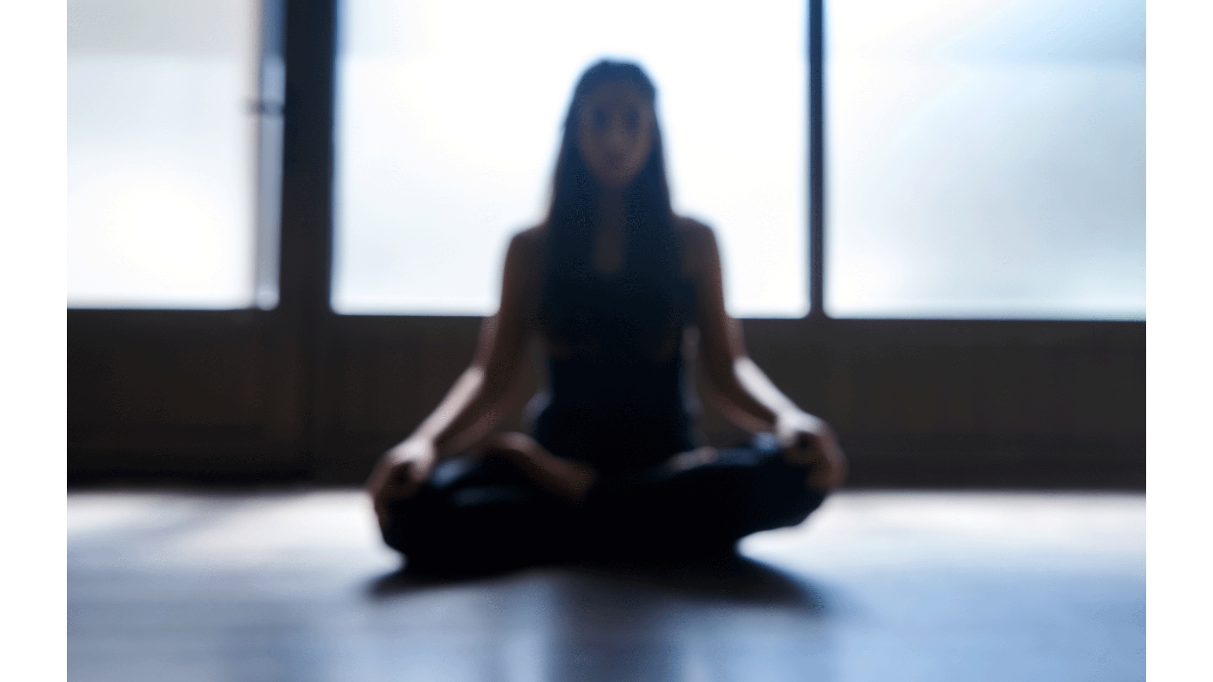 A woman sitting cross-legged in a dark room with bright windows behind her.
