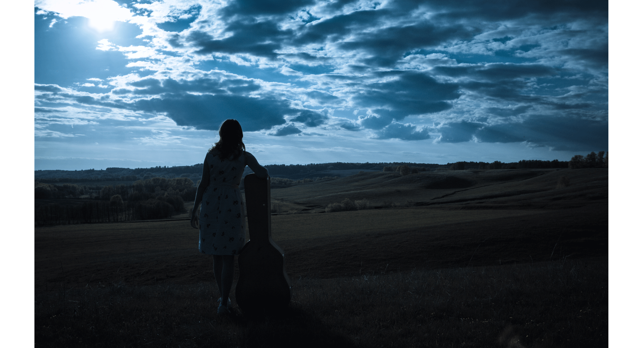 Woman standing in an open field beneath the full Moon in Capricorn while holding a guitar case