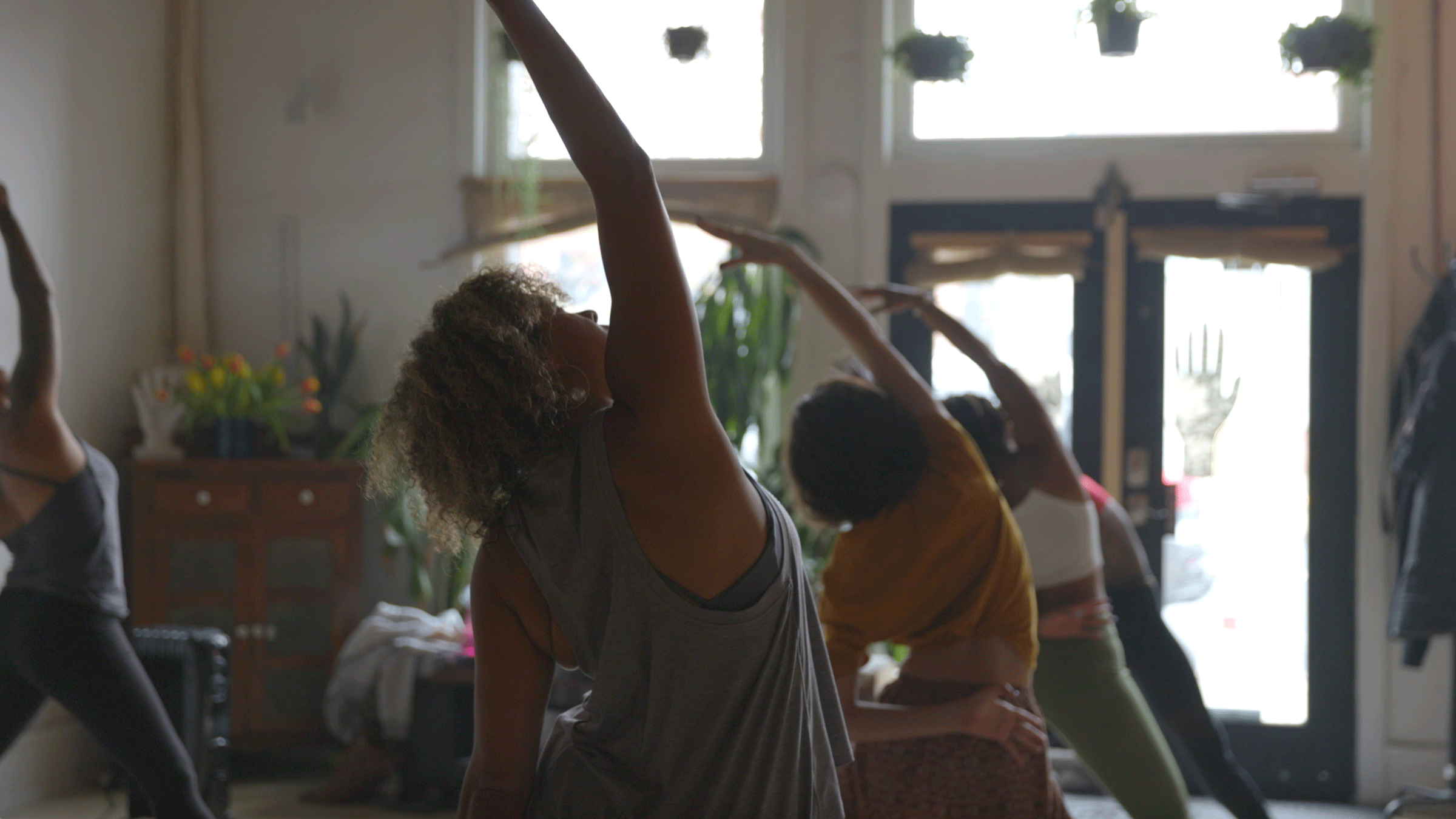 Women in a yoga class with hardwood floors and windows in the background while the teacher takes them through yoga sequences