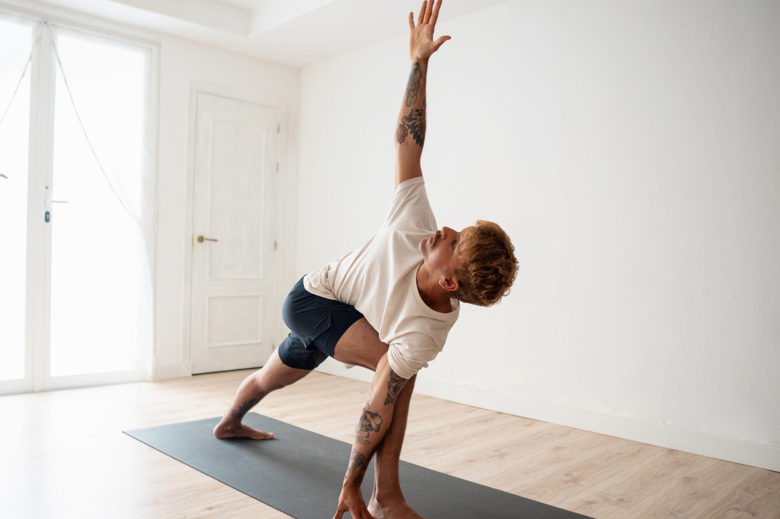 Man in revolved lunge on yoga mat on wooden floor with surrounded by white walls with light streaming in windows.