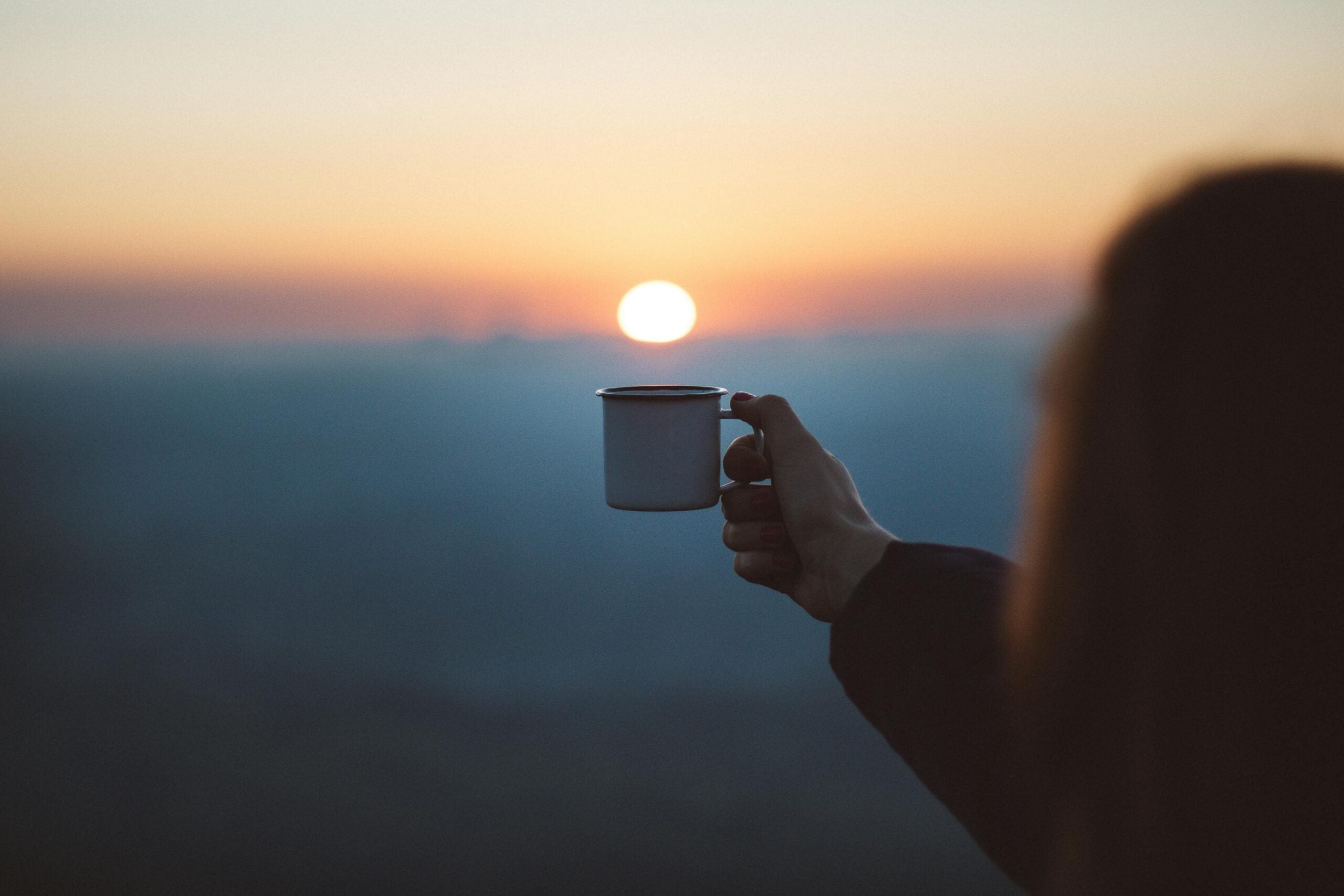 Woman holding cup of tea in front of an orange sunset.