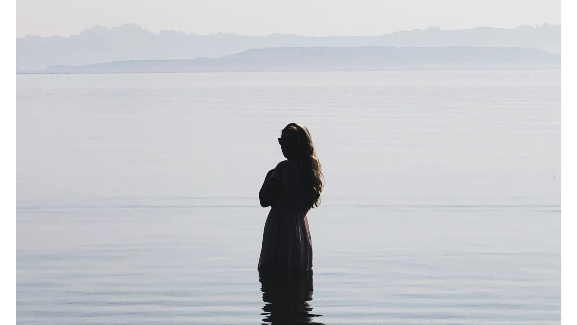 Woman in a dress standing in a sea that's smooth just before sunset with clouds